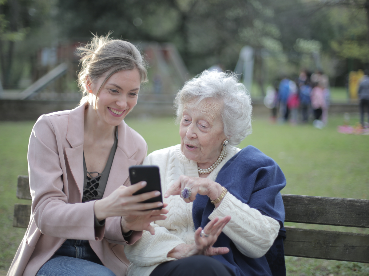 A younger woman and an older woman sitting together on a bench, looking at a smartphone. This symbolizes the significance of National Home Care and Hospice Month