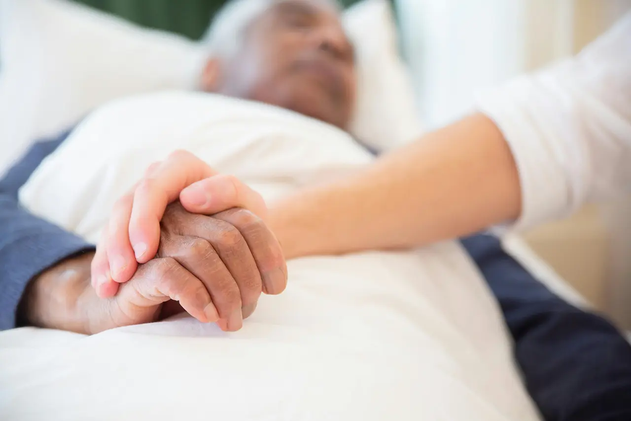 elderly man laying in a bed holding the hand of a caretaker